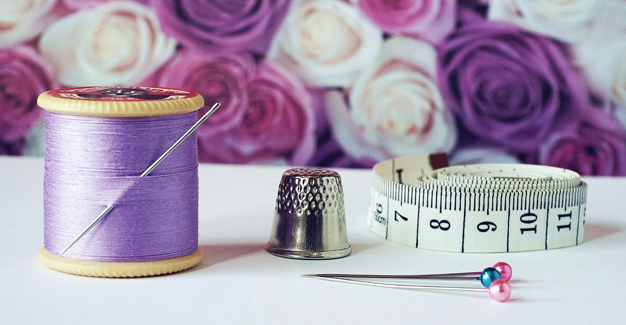 A close-up of sewing tools including lilac thread, a thimble, tape measure, and colorful pins with a rose backdrop.